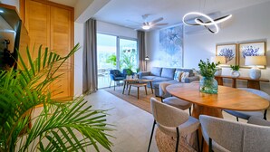 A spacious living area with a wooden dining table, comfortable seating, and a large window offering a view of the outdoors. - Cana Brava Residences - Elite Apartment, Golf View (Punta Cana)