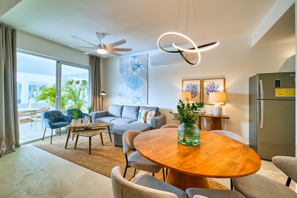 A modern living room with a round wooden dining table, grey sofas, and a ceiling fan.