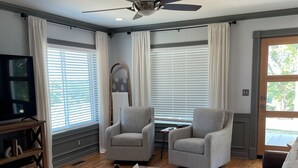 Un salon spacieux avec une table basse en bois, deux fauteuils gris, un canapé en cuir marron, un ventilateur de plafond et un téléviseur posé sur un meuble en bois.