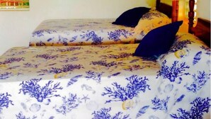 Una habitación con dos camas, una ventana con persiana verde estampada, un espejo con marco azul y una lámpara amarilla.