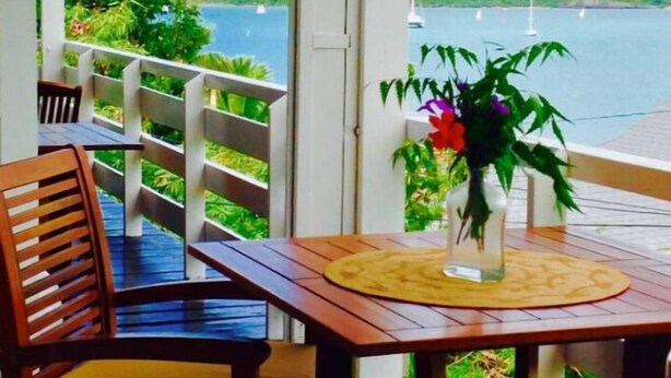 Una mesa de madera con sillas y un florero en un balcón con vista a un cuerpo de agua y colinas verdes.