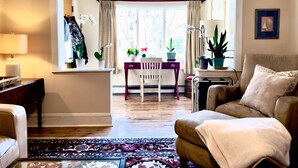 A living room with a sofa, a chair, a small table with plants, and a view through a window.