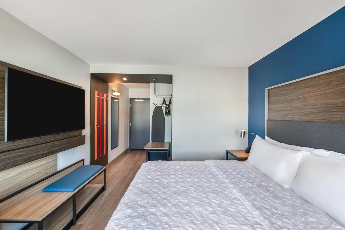 A modern hotel room with a large bed, a TV, a bench, a desk, and a wardrobe.