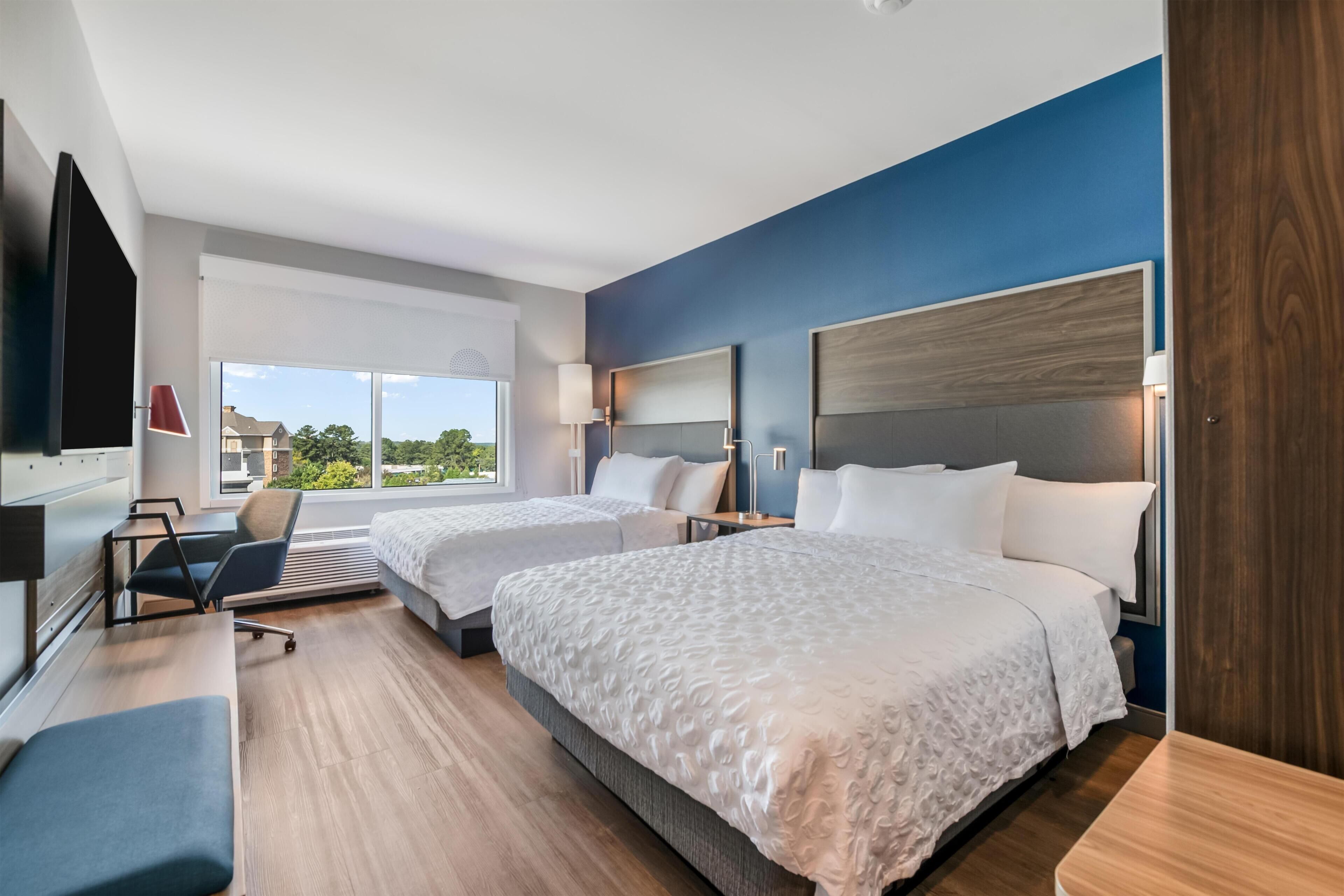 A hotel room with two beds, a desk, a chair, a TV, and a large window with a view.