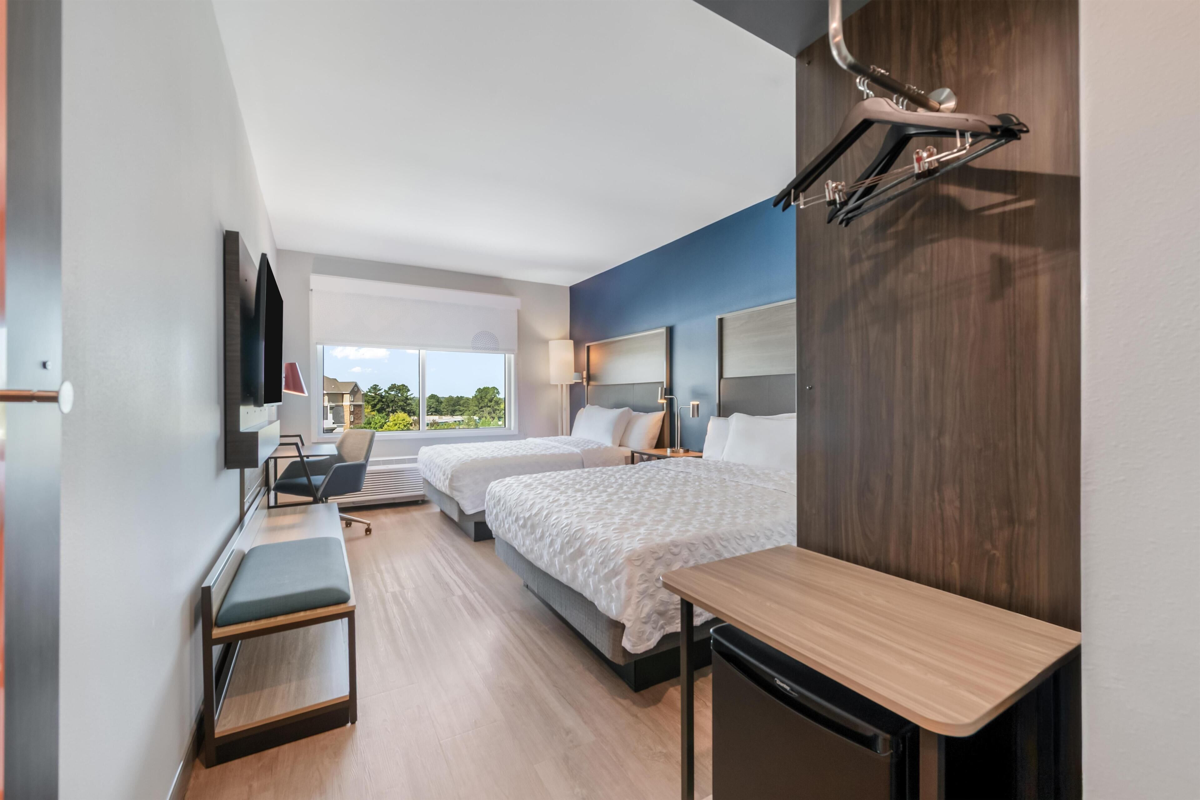 A modern hotel room with two beds, a desk, a bench, a TV, and a window with a view of greenery.