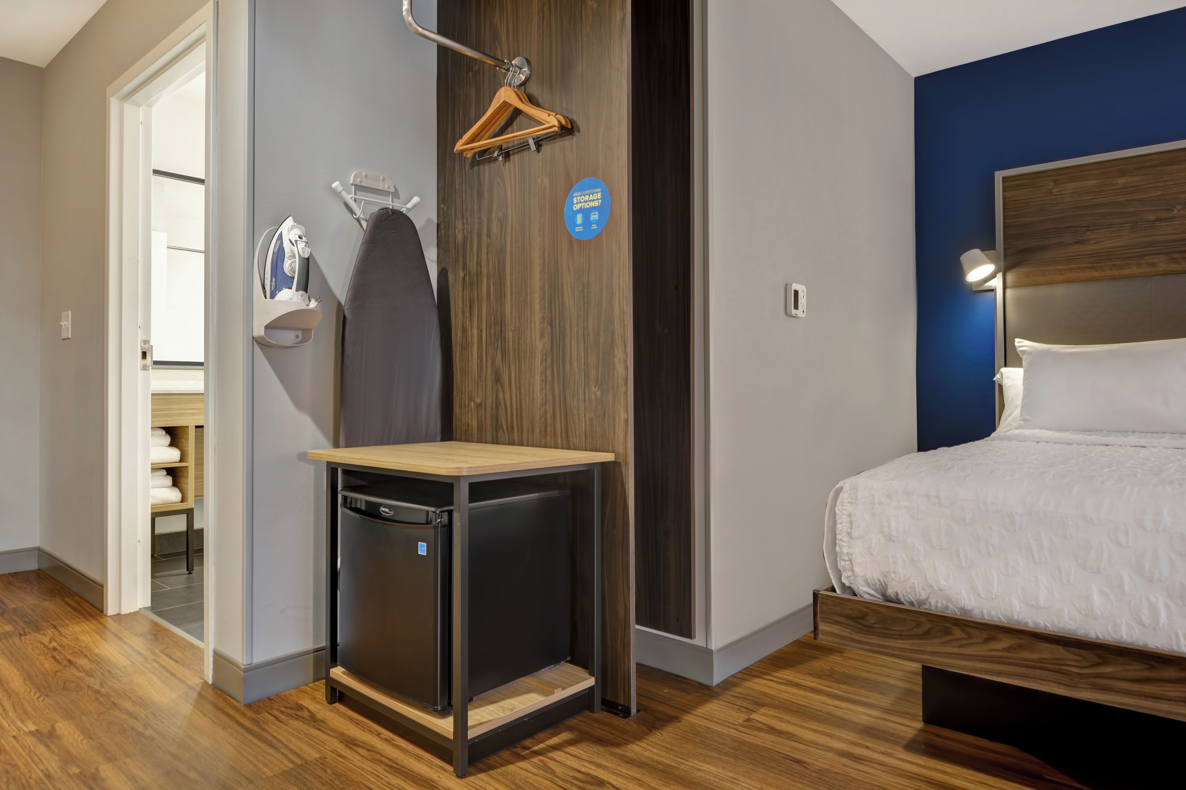 Habitación de hotel pequeña con cama, una mesita, un calentador de pared, un letrero de almacenamiento y piso de madera.