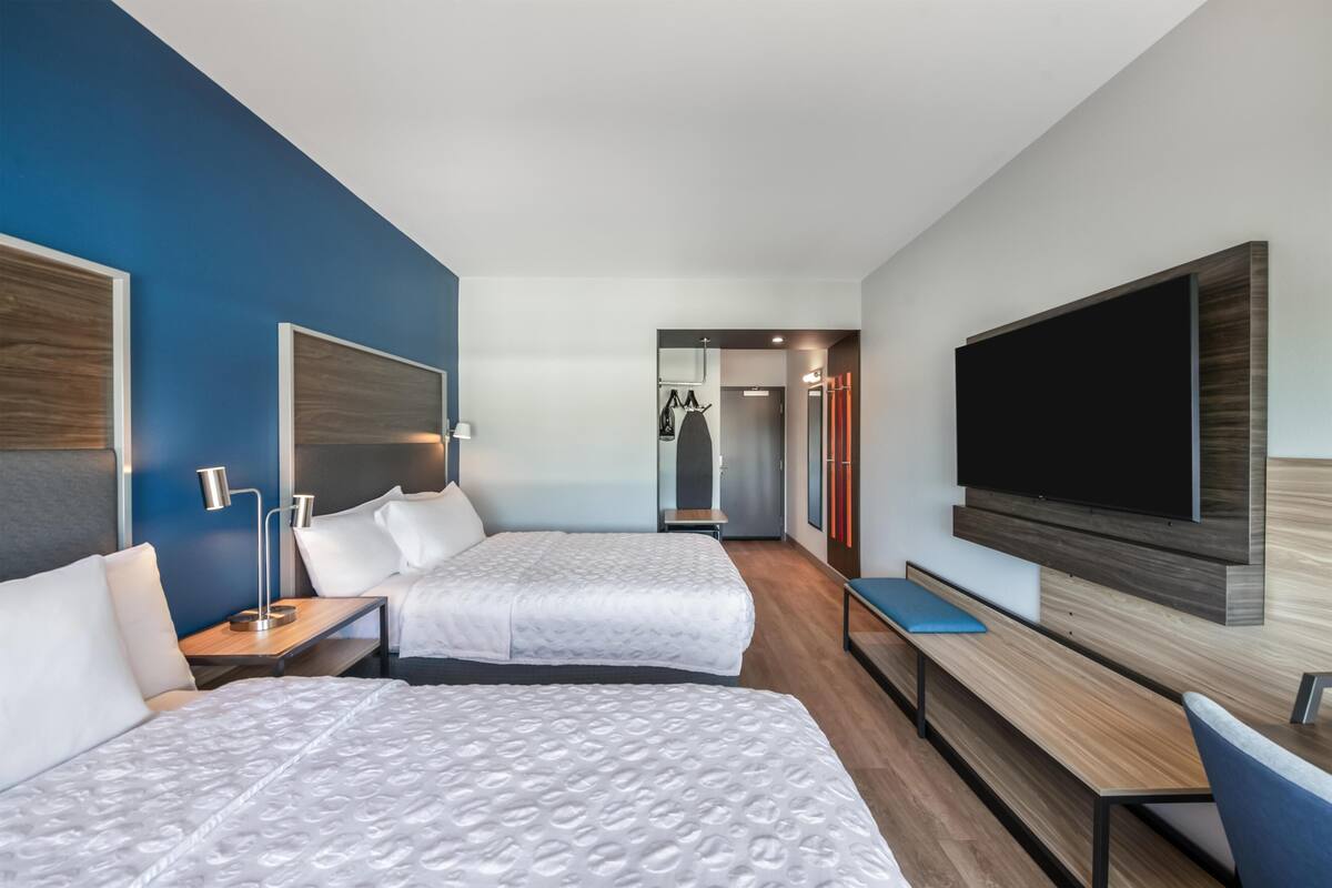 A hotel room with two beds, a TV, a desk, and a bench.