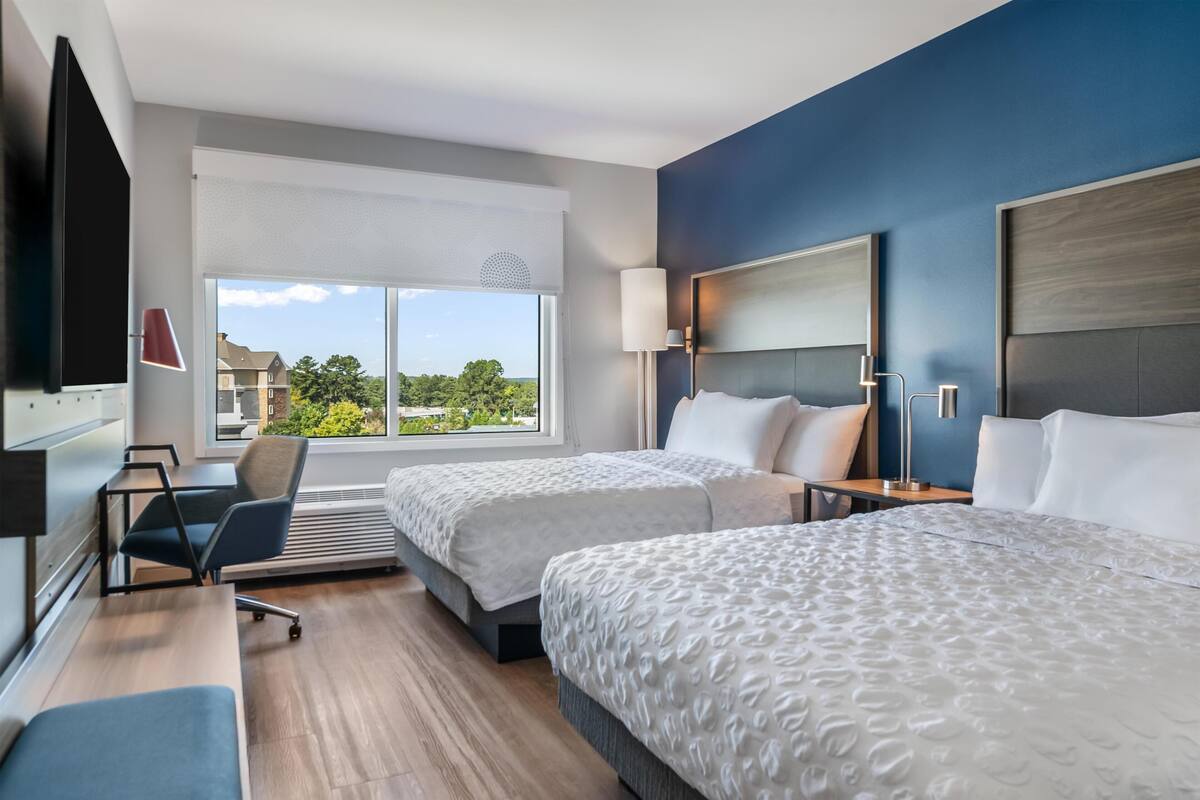 A hotel room with two beds, a desk, a chair, a TV, and a large window with a view of buildings and trees.