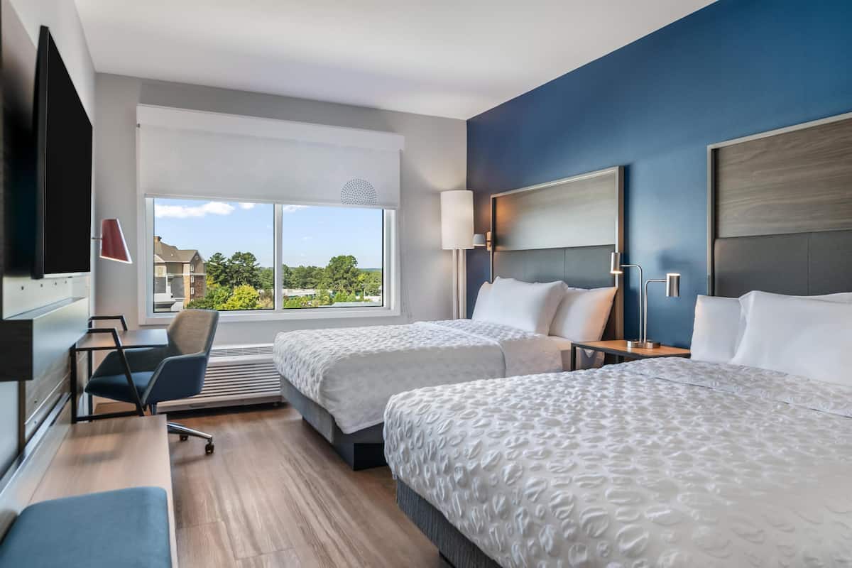 Habitación de hotel con dos camas, un escritorio, una silla, un televisor y un amplio ventanal con vistas a edificios y árboles.