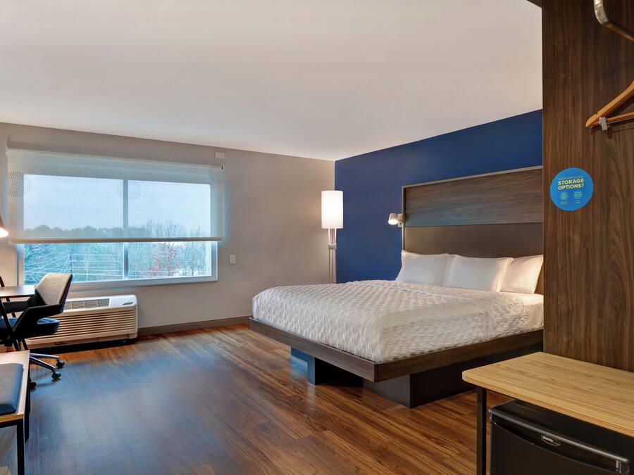 A hotel room with a bed, a desk, a chair, a window, and a wall-mounted heater.