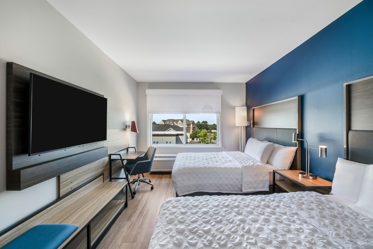 A hotel room with a TV, two beds, a desk, and a window with a city view.