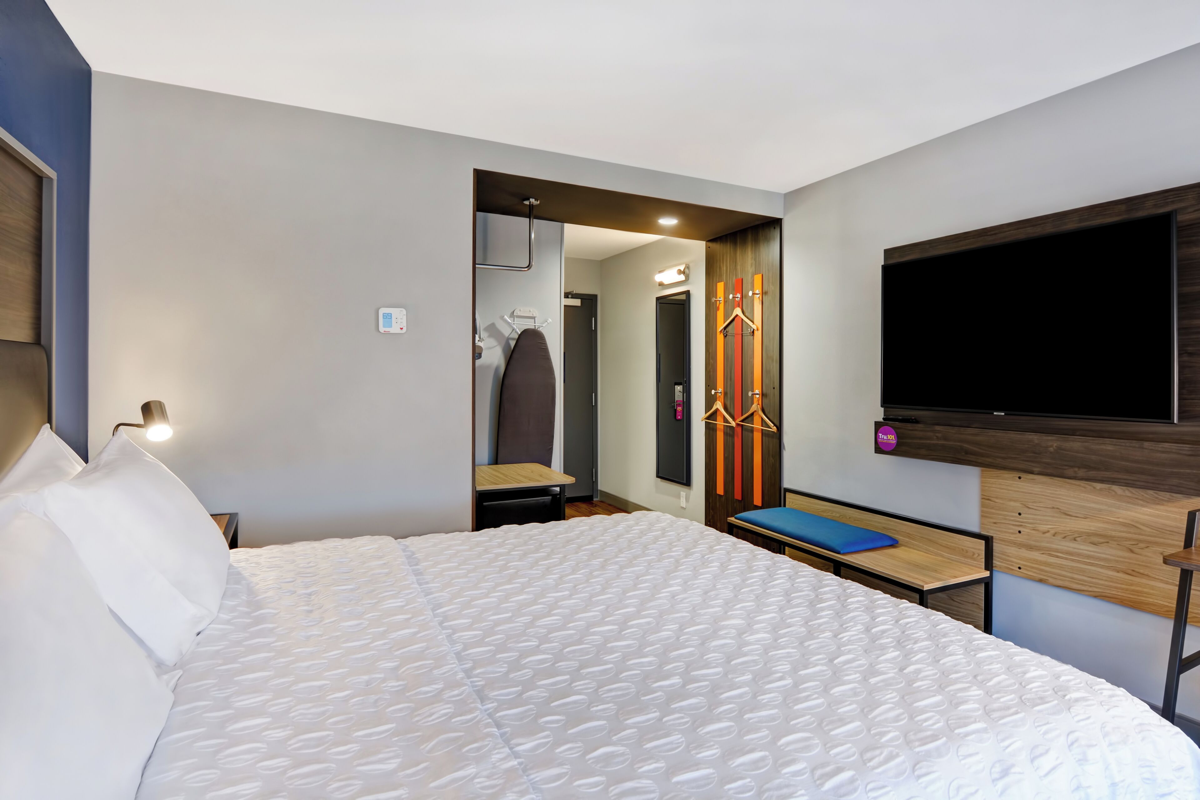 A modern hotel room with a large bed, a TV mounted on the wall, a seating area with a blue cushion, and a mirror with a wooden frame.