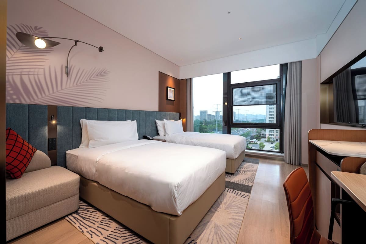 Een moderne hotelkamer met twee bedden, een bank, een bureau en een groot raam met uitzicht op de stad.