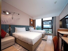 Una habitación de hotel moderna con dos camas, un sofá, un escritorio y un amplio ventanal con vista a la ciudad.