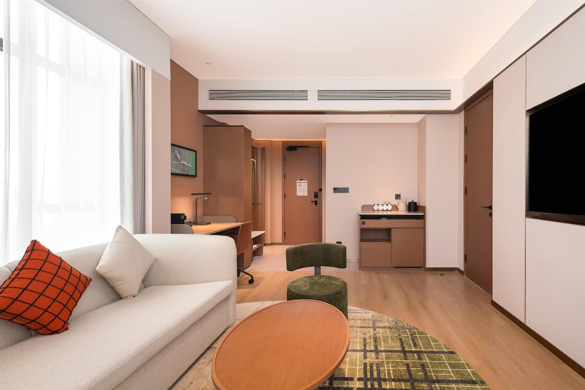 Een moderne hotelkamer met een bank, een klein rond tafeltje, een groene poef, een kitchenette en een groot raam.