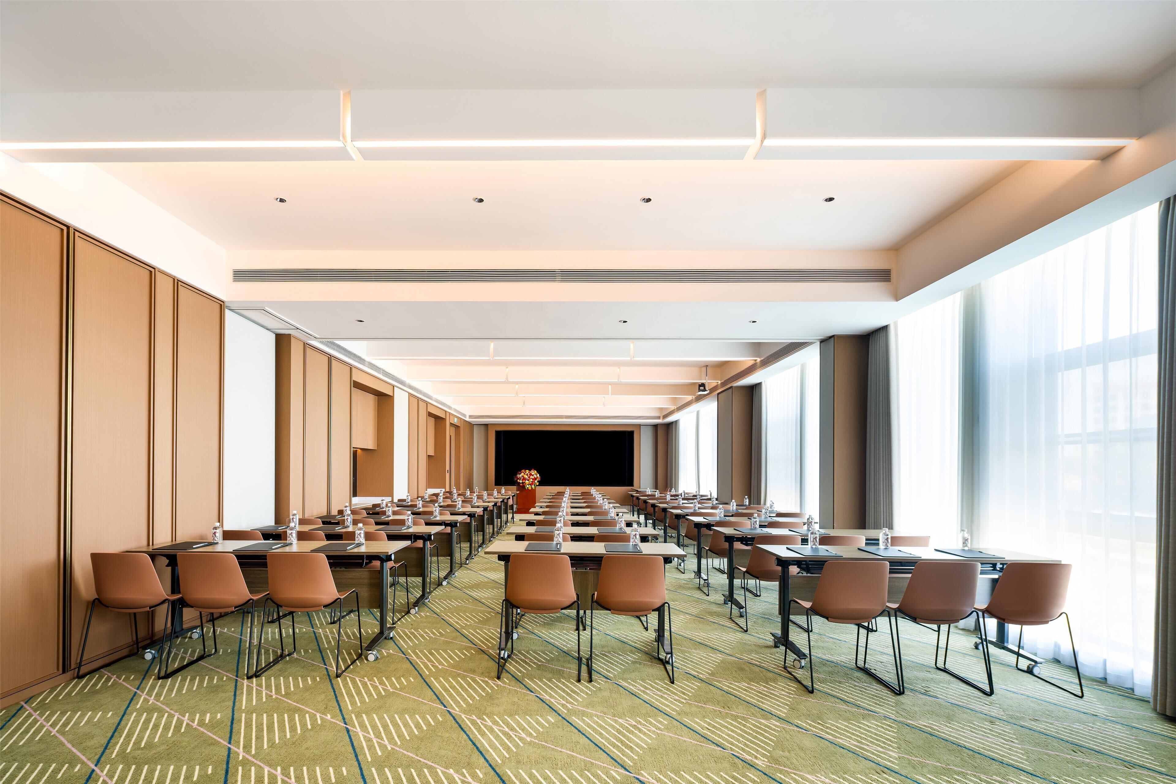 A conference room with rows of tables and chairs, large windows, and a stage at the far end.