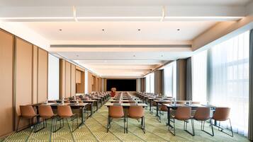 A conference room with rows of tables and chairs, large windows, and a stage at the far end.