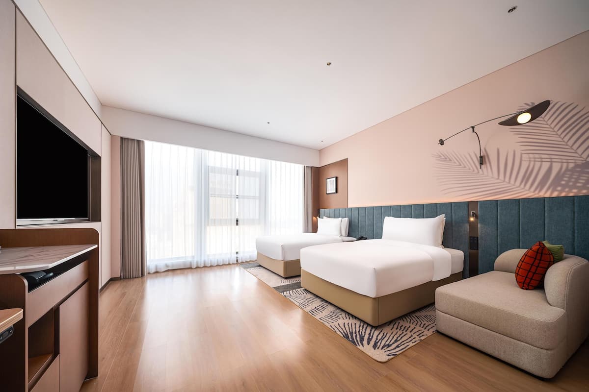 Een moderne hotelkamer met een grote flatscreen televisie, twee bedden, een bank en een groot raam met dunne gordijnen.