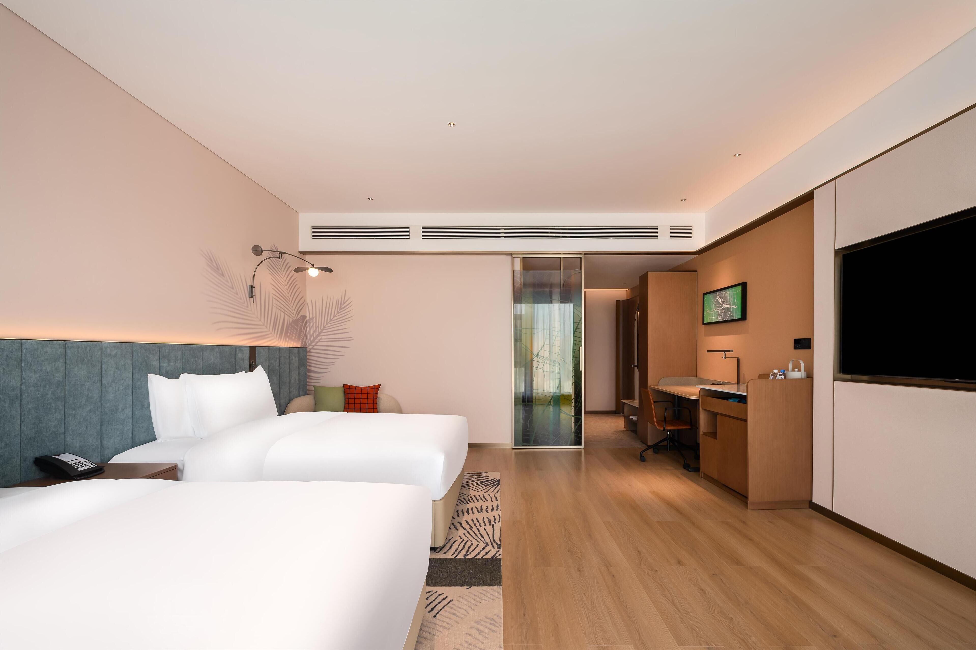 Ein modernes Hotelzimmer mit zwei Betten, einem Schreibtisch, einem Fernseher und einer Schiebetür zu einem Badezimmer.