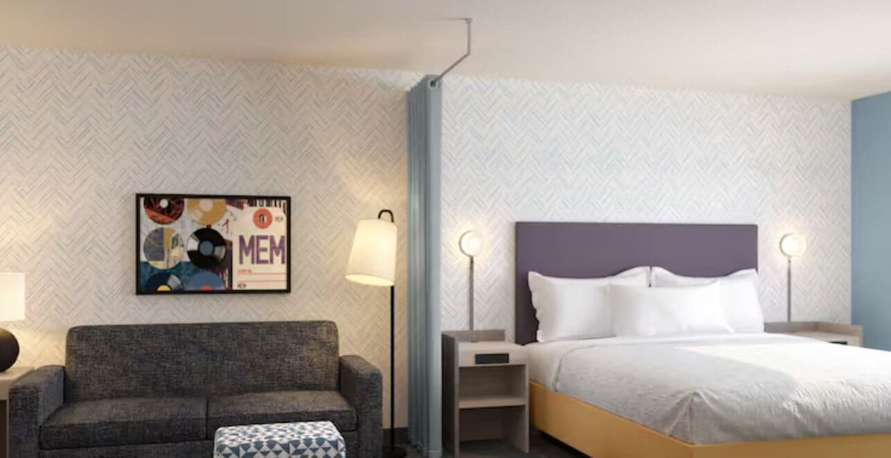 A hotel room with a bed, a sofa, a side table, a lamp, and a wall art piece.