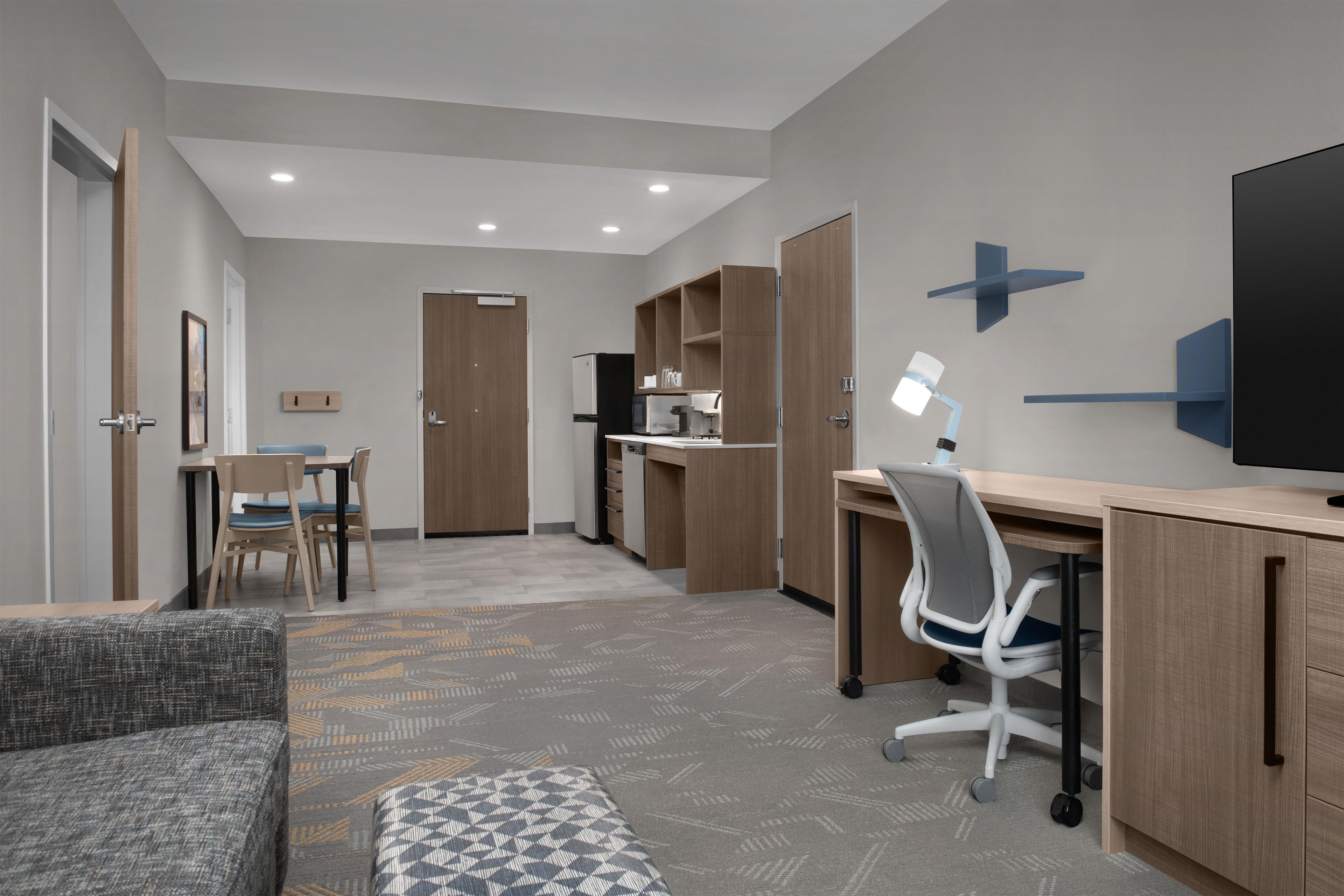 A modern hotel room with a dining area, kitchenette, and a large flat-screen TV mounted on the wall.