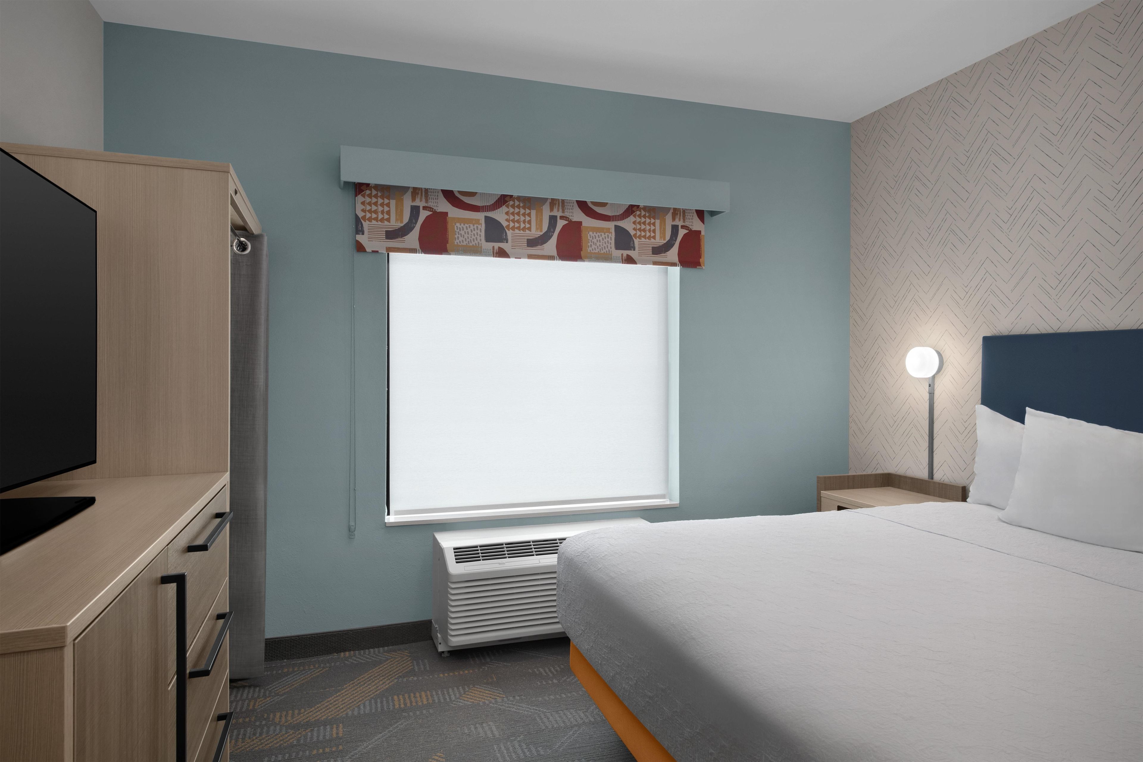 Una habitación de hotel con cama, televisión, armario, ventana con cortina estampada, mesita de noche con lámpara y pared decorada.