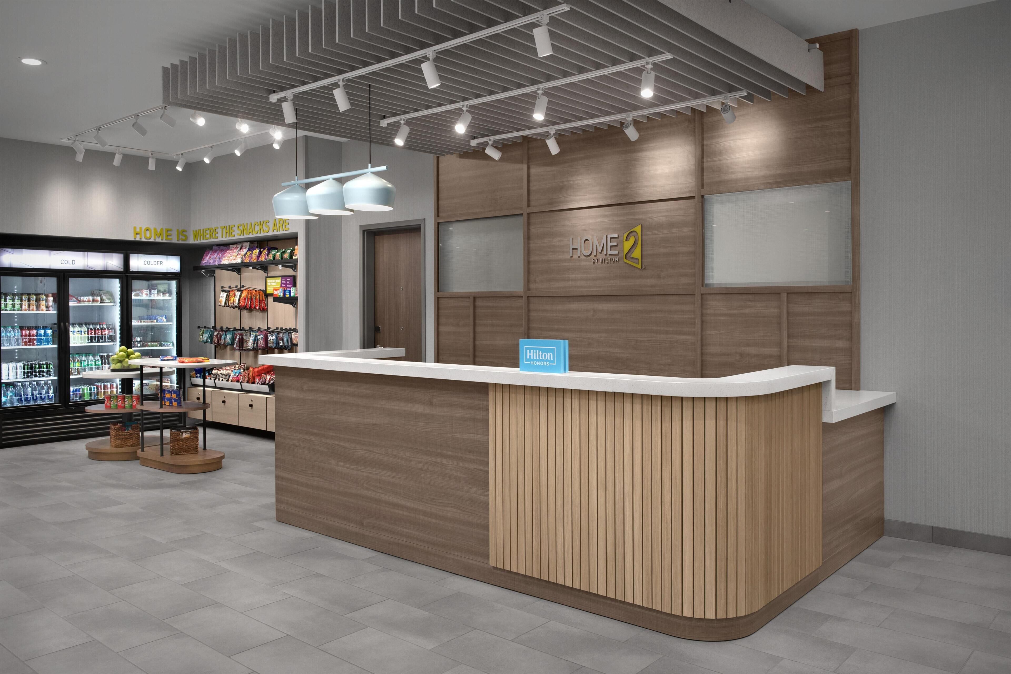 A modern hotel reception area with a wooden reception desk, a 'Home 2' sign, a 'Hilton' sign, and a refrigerator stocked with snacks.