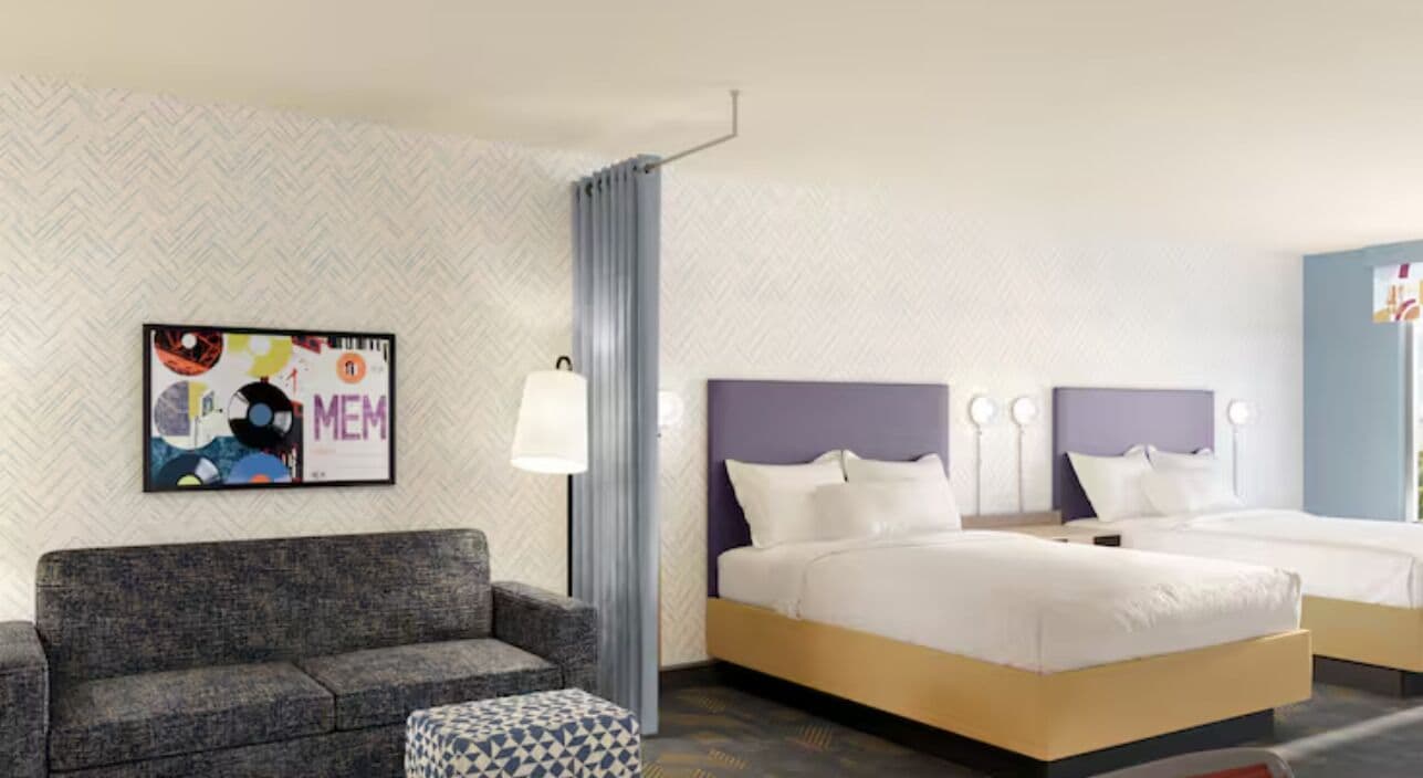 A hotel room with a bed, a sofa, a side table, and a wall-mounted artwork.