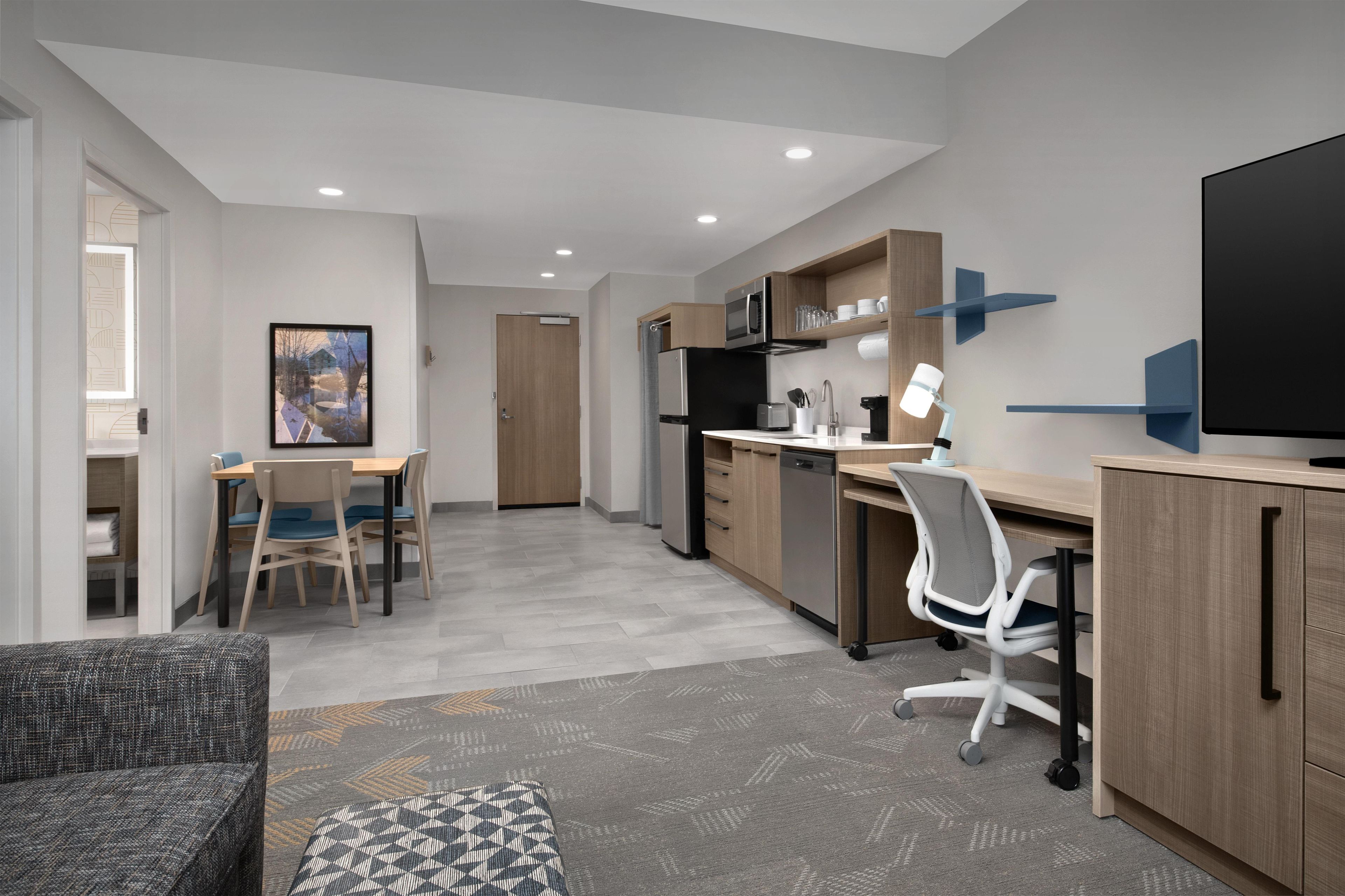 Habitación moderna y bien iluminada con zona de comedor, cocina y sala de estar con sofá y reposapiés estampado.
