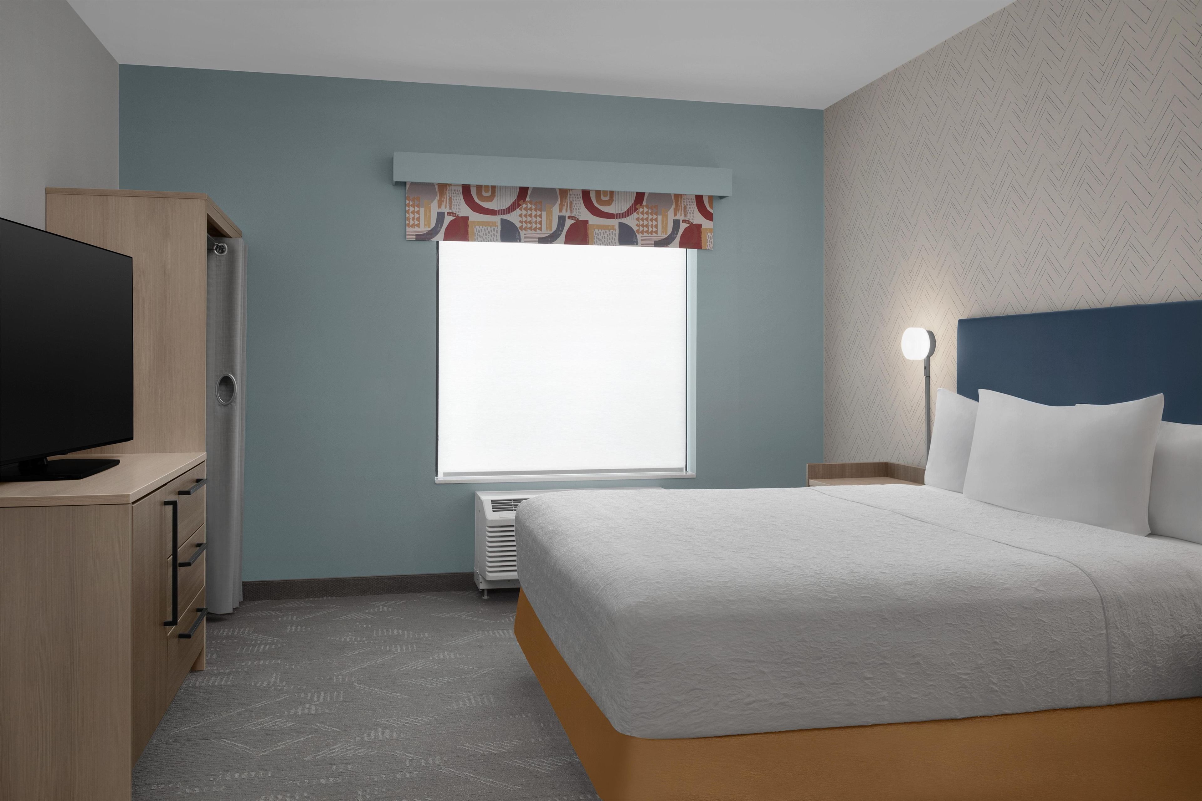 Una habitación de hotel con cama, televisor, armario y ventana con cortina estampada.