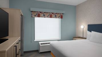 A hotel room with a bed, a TV, a heater, a window with a patterned valance, and a nightstand with a lamp.