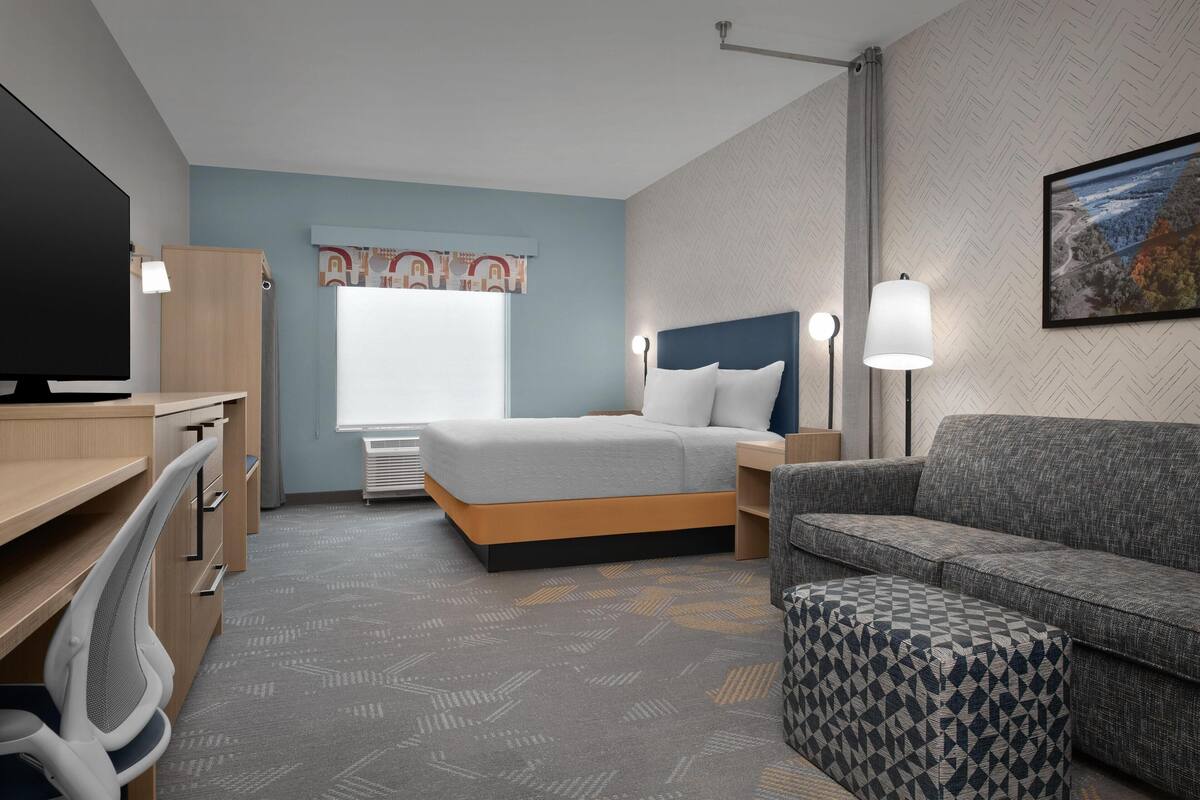 Una habitación de hotel con cama, escritorio, silla, sofá, lámpara y un cuadro en la pared.