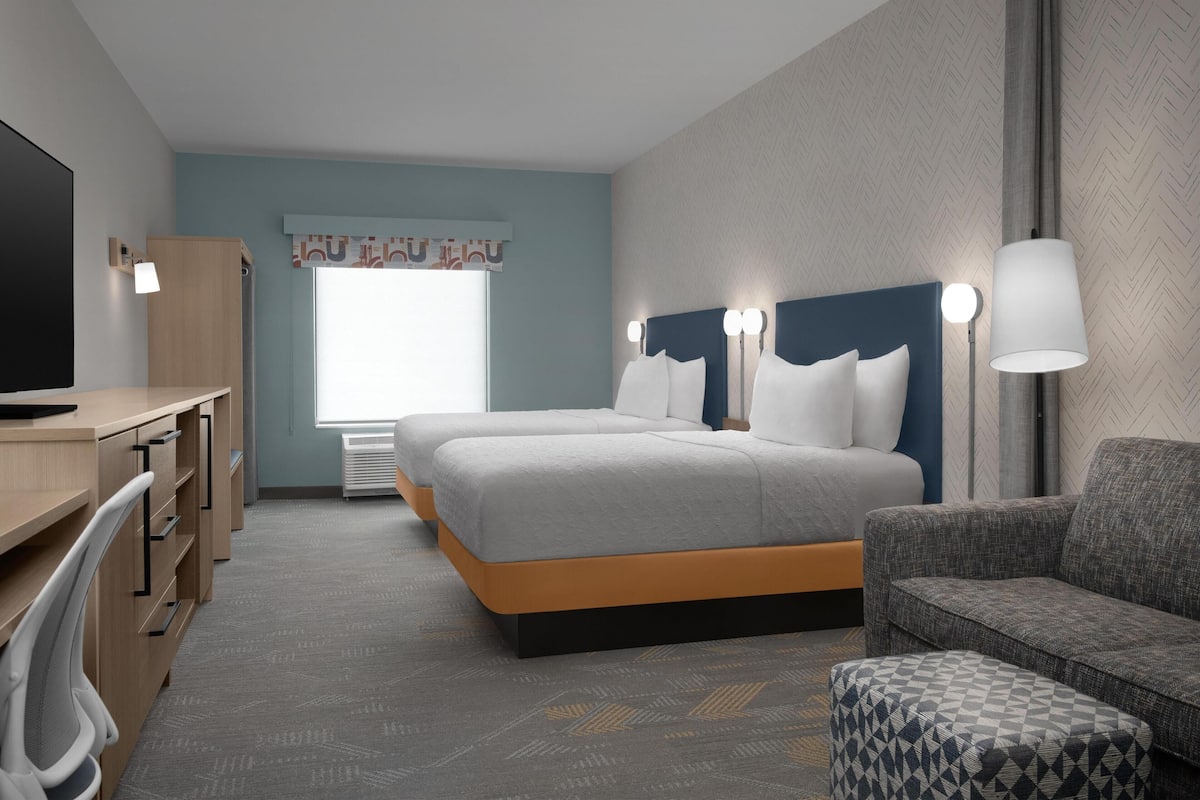 Una habitación de hotel con escritorio, dos camas, un sofá y un otoman con estampado.