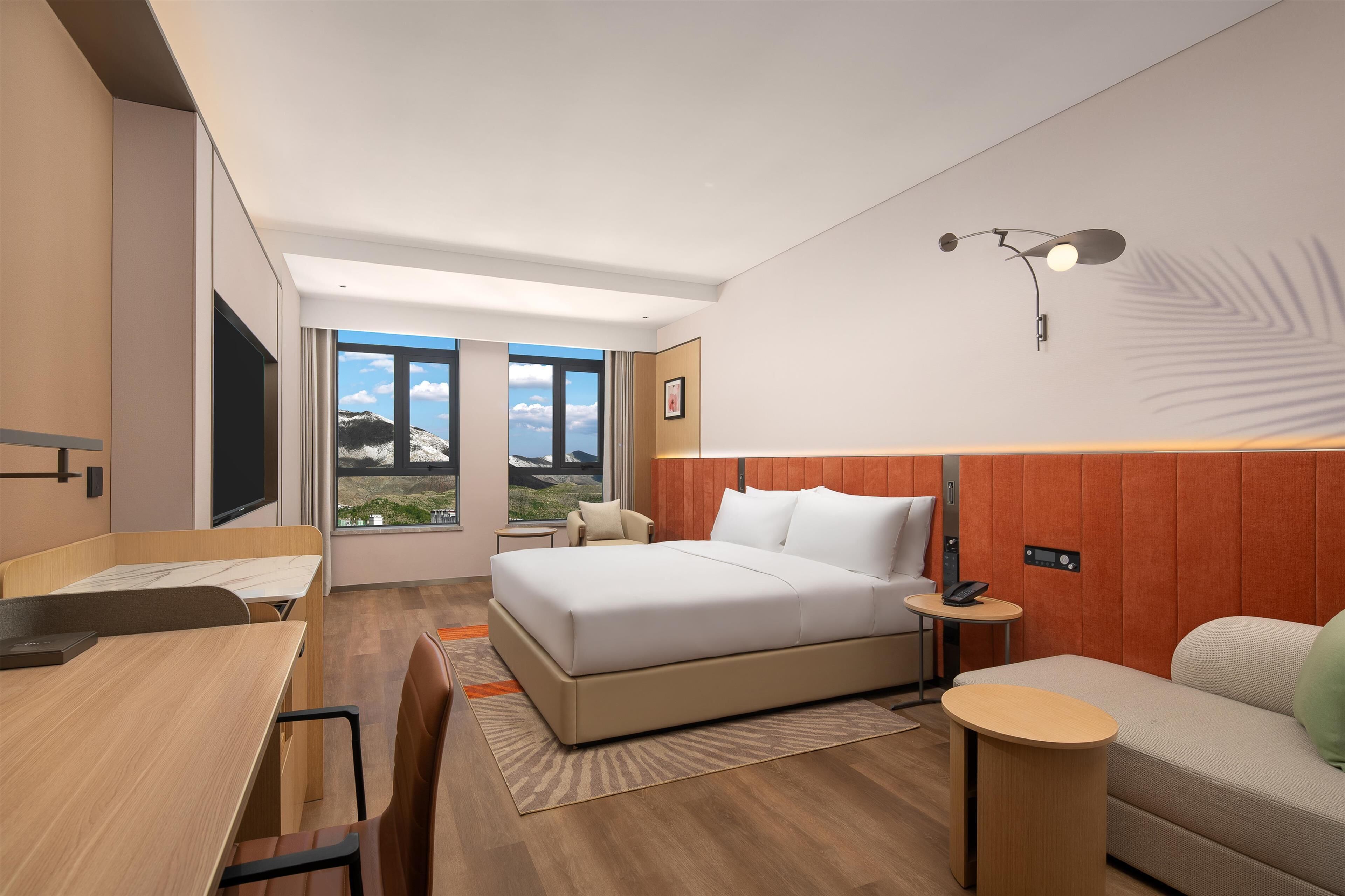A modern hotel room with a large bed, a desk, a sofa, and a view of the outdoors.