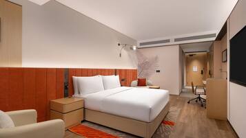 A modern hotel room with a large bed, a desk, and a chair. There is a nightstand with a lamp, a wall-mounted TV, and a decorative plant.