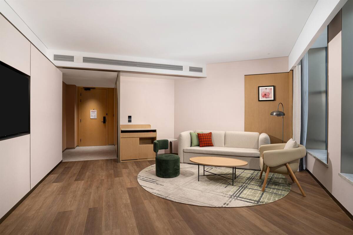 Een moderne hotelkamer met een bank, een klein tafeltje, een groene poef, een bureau en een televisie.