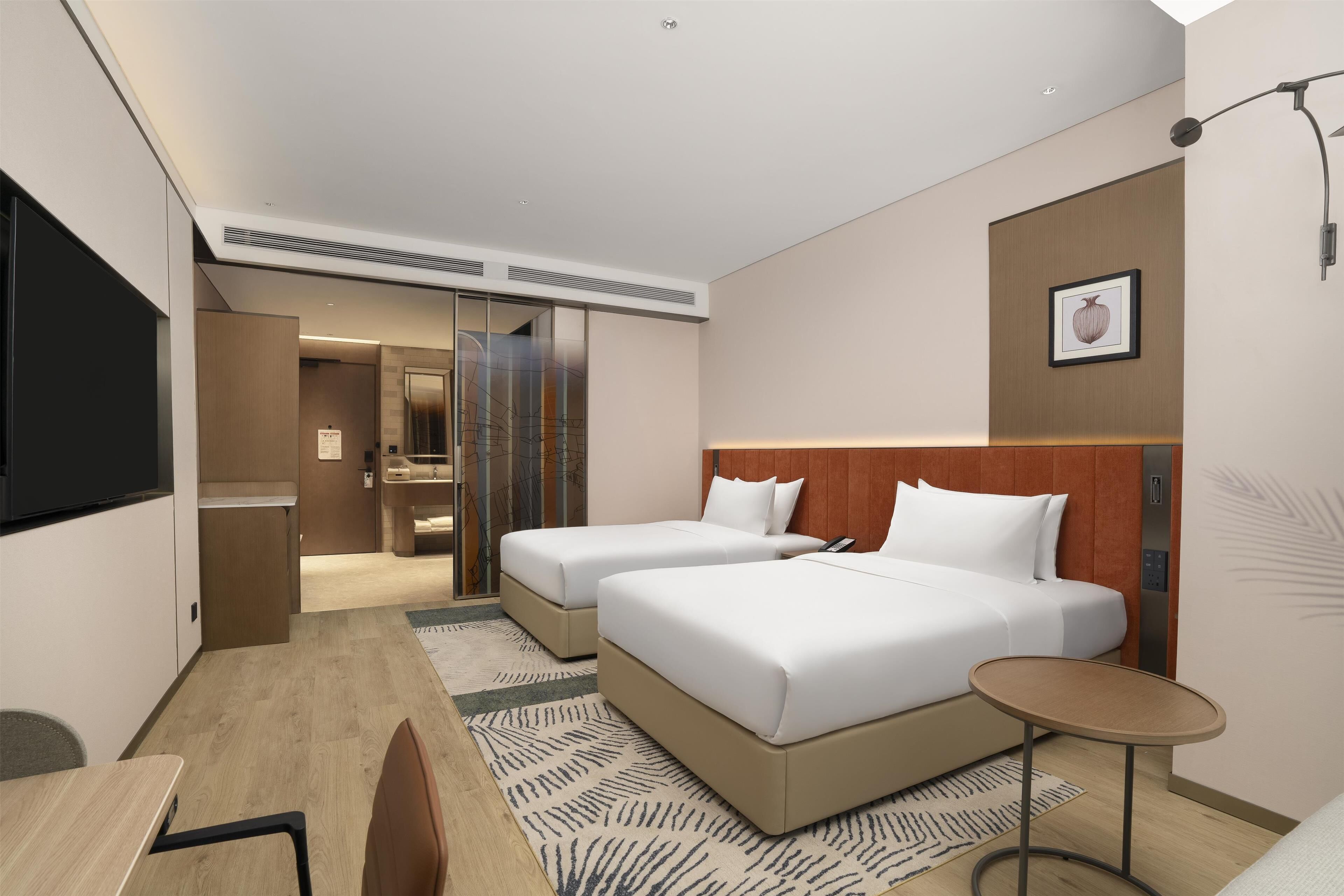 A modern hotel room with two beds, a desk, a chair, a small table, and a TV.