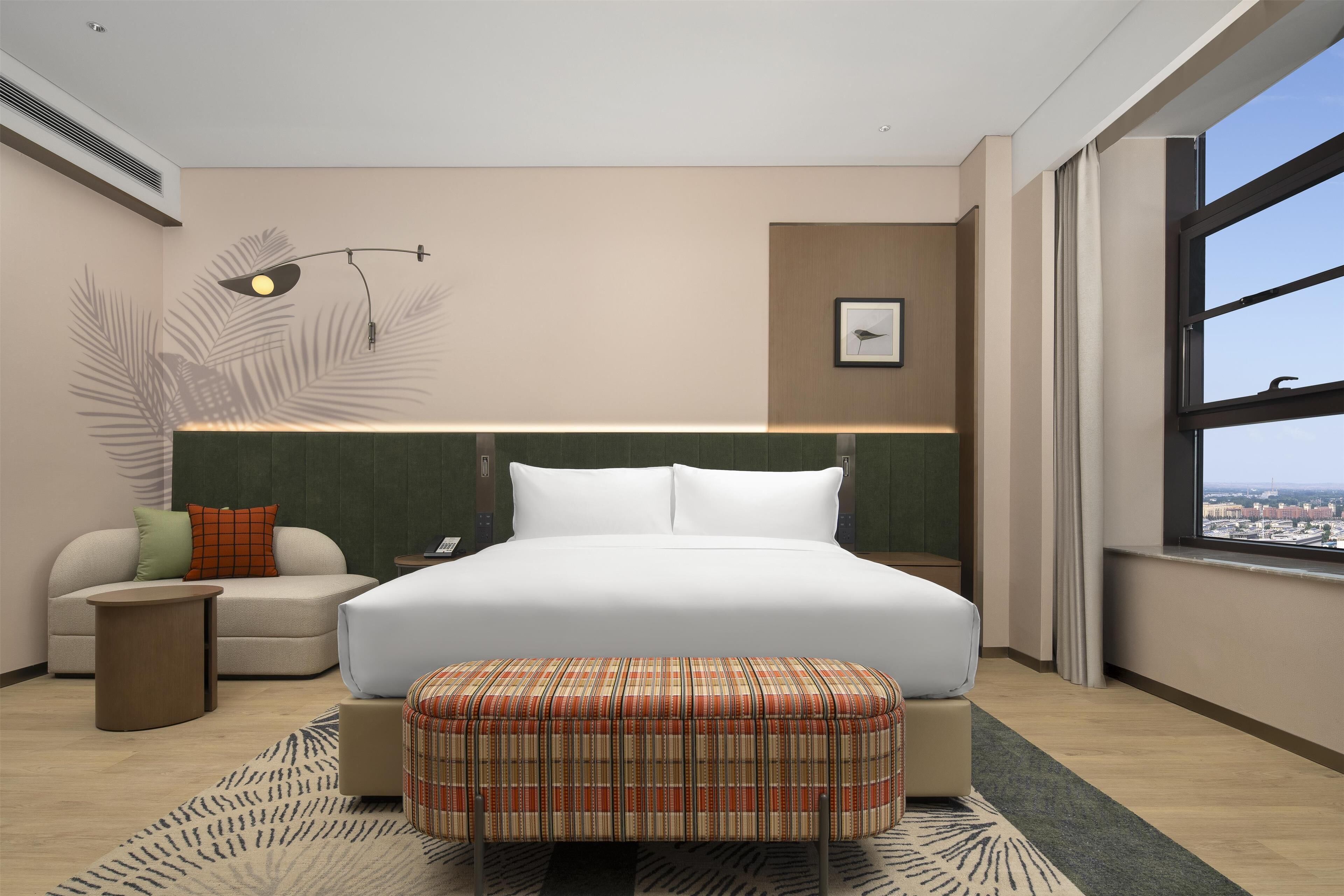 Habitación de hotel moderna con una cama grande, un sofá, una mesita auxiliar y vistas a la ciudad a través de un ventanal.