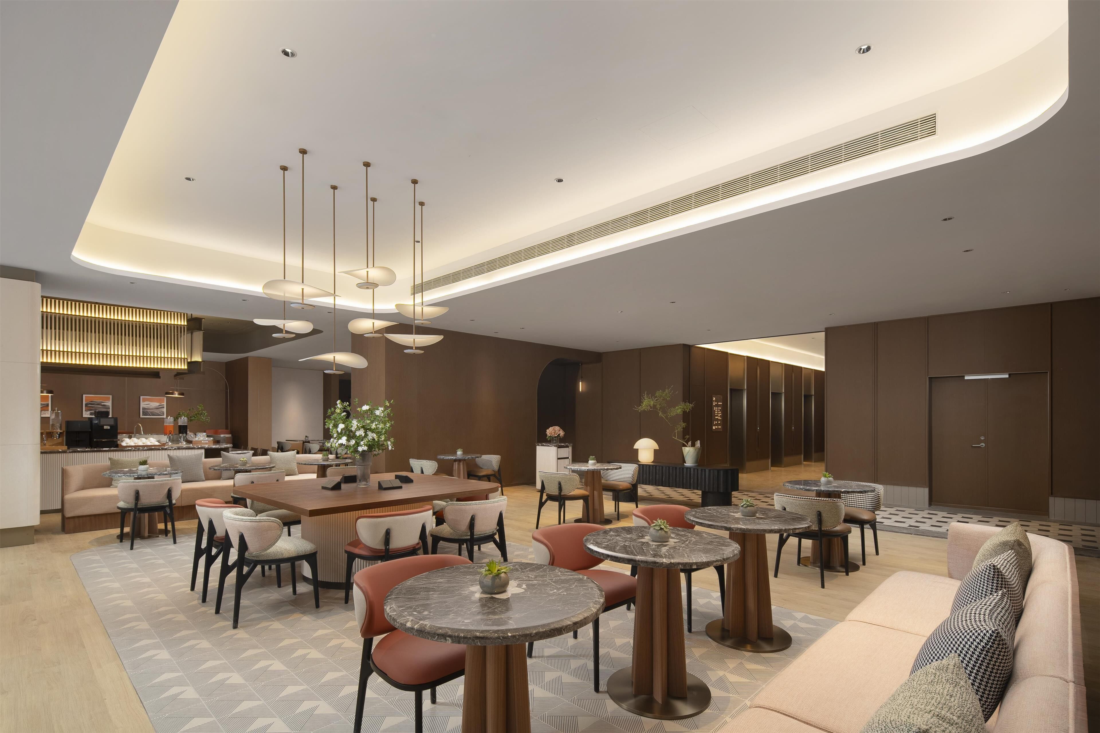 A modern hotel lobby with a central bar area, multiple seating arrangements including round tables and sofas, and a decorative ceiling with integrated lighting.