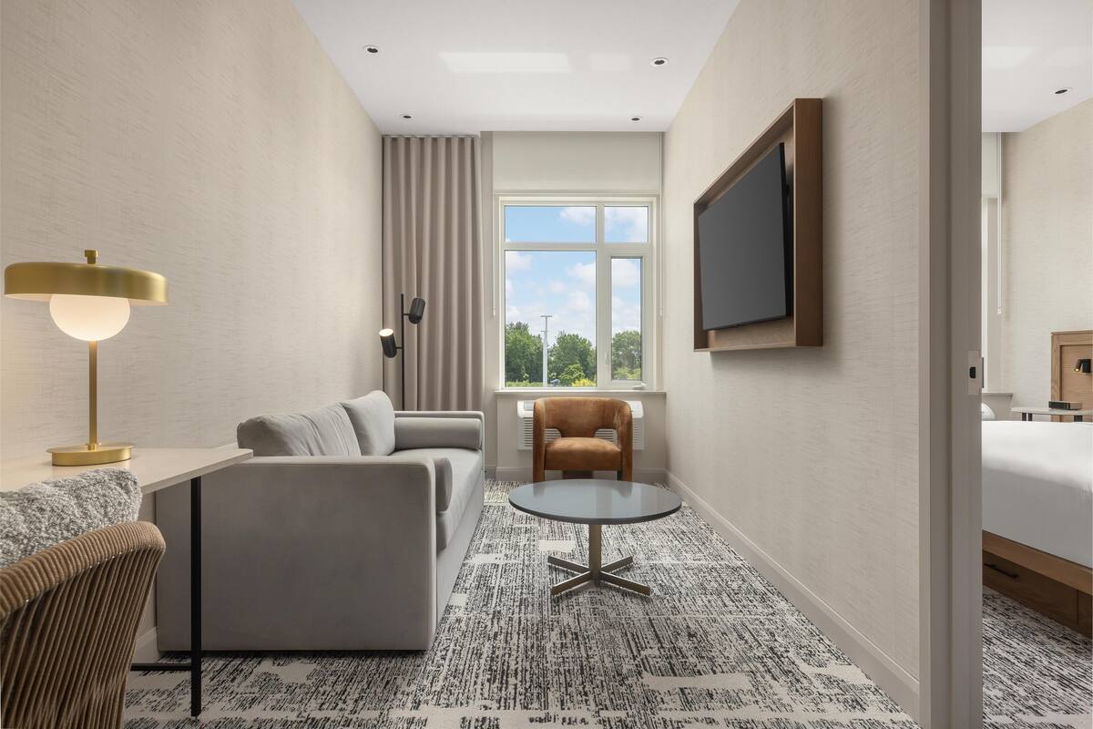 Et moderne hotelværelse med sofa, rundt bord, fjernsyn ophængt på væggen og et vindue med udsigt til træer.