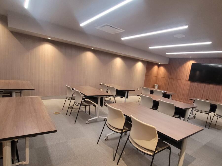 Uma sala de conferências com várias mesas e cadeiras, iluminação embutida no teto e uma TV de tela plana fixada na parede.