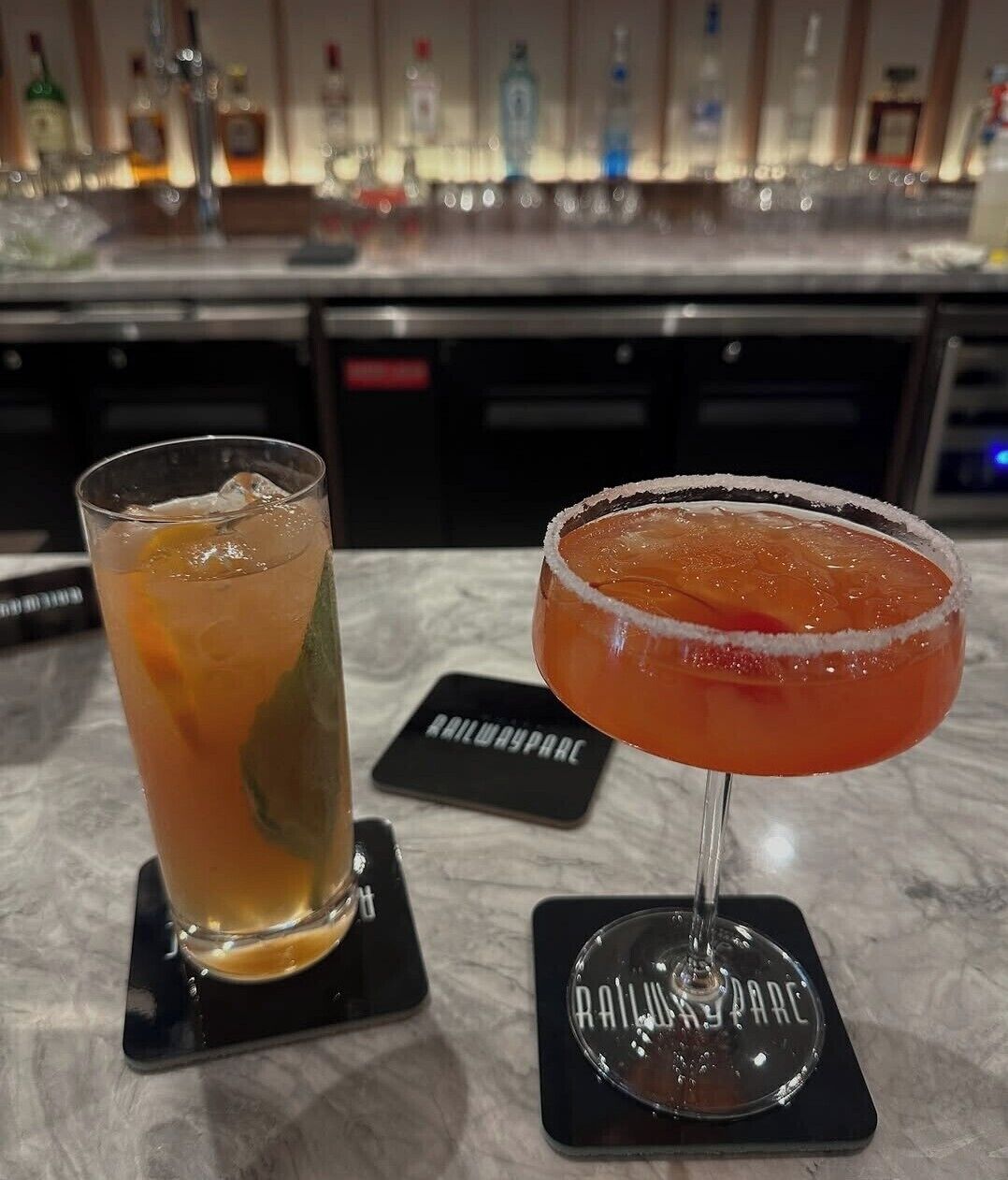 A bar area with a glass of iced drink and a margarita on a coaster, with a bar counter and bottles in the background.