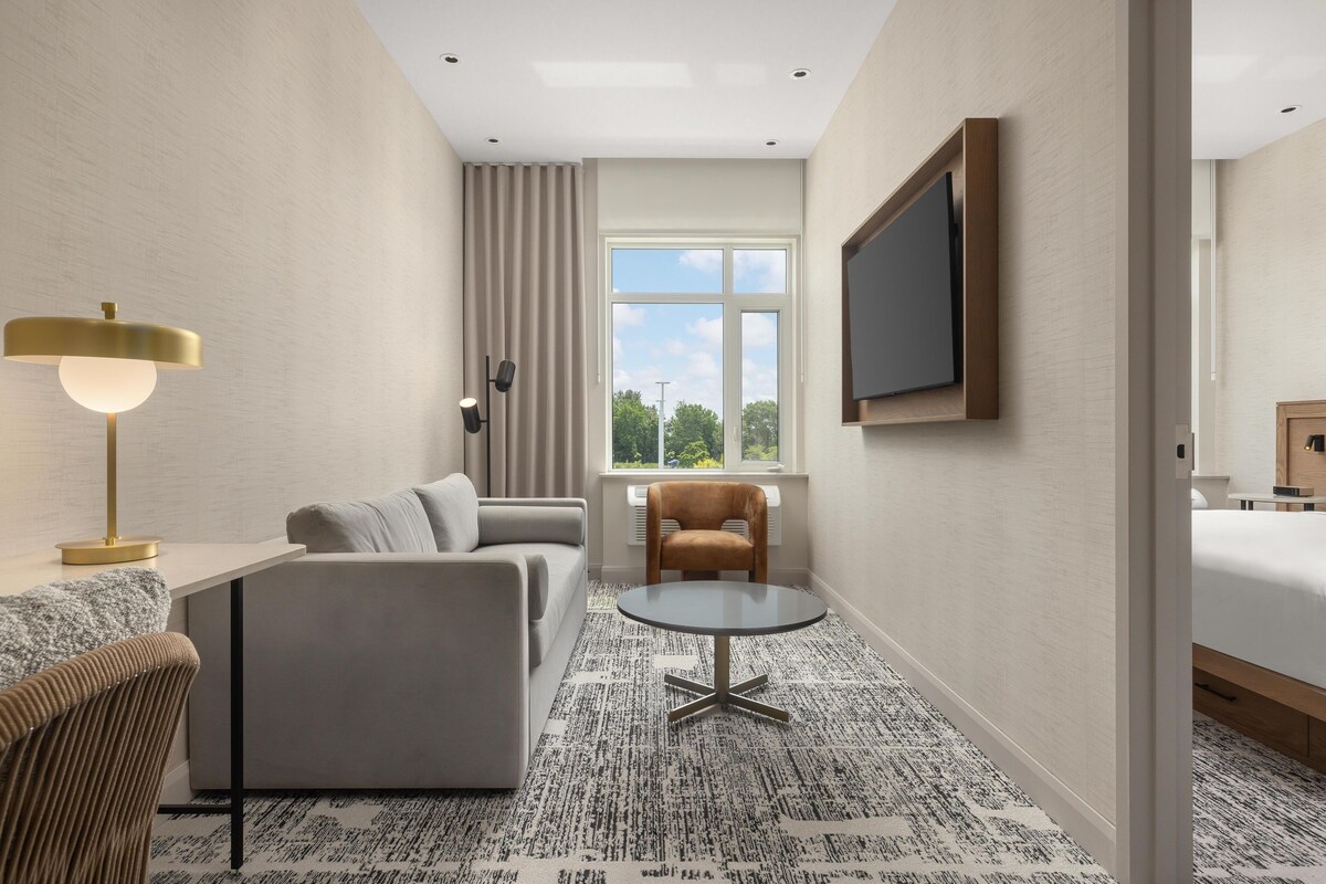 Een moderne hotelkamer met een bank, een ronde tafel, een aan de muur bevestigde televisie en een raam met uitzicht op bomen.