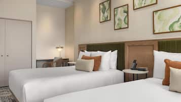 A hotel room with two beds, a desk, a chair, and a small table with a phone and a charging device.
