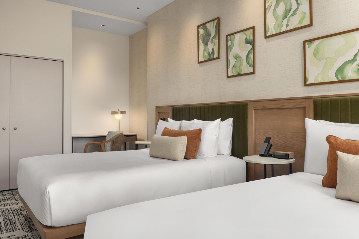 A hotel room with two beds, a desk, a chair, a nightstand with a phone, and artwork on the wall.