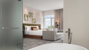 A modern hotel room with a large bed, a sofa, a bathtub, and a large window with curtains. - Hotel Railwayparc Montreal, Tapestry Collection By Hilton (Montreal)
