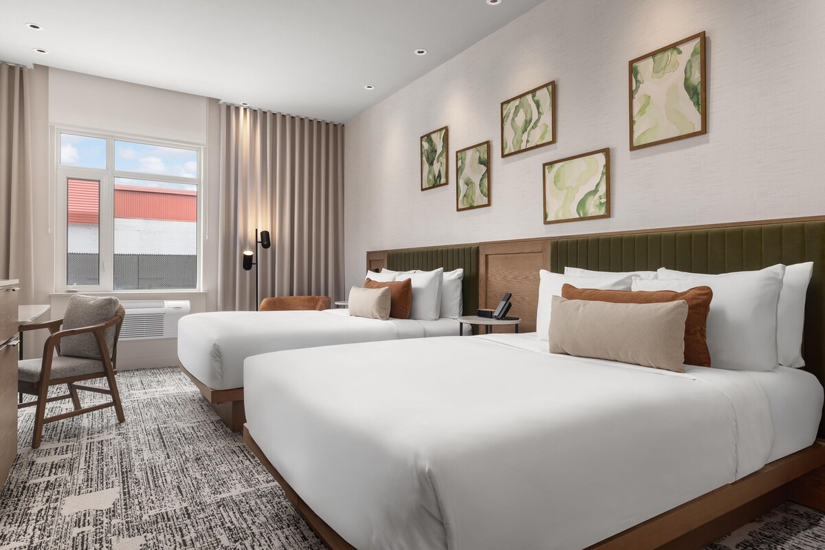A hotel room with two beds, a desk, and a chair. There are four framed pictures on the wall.