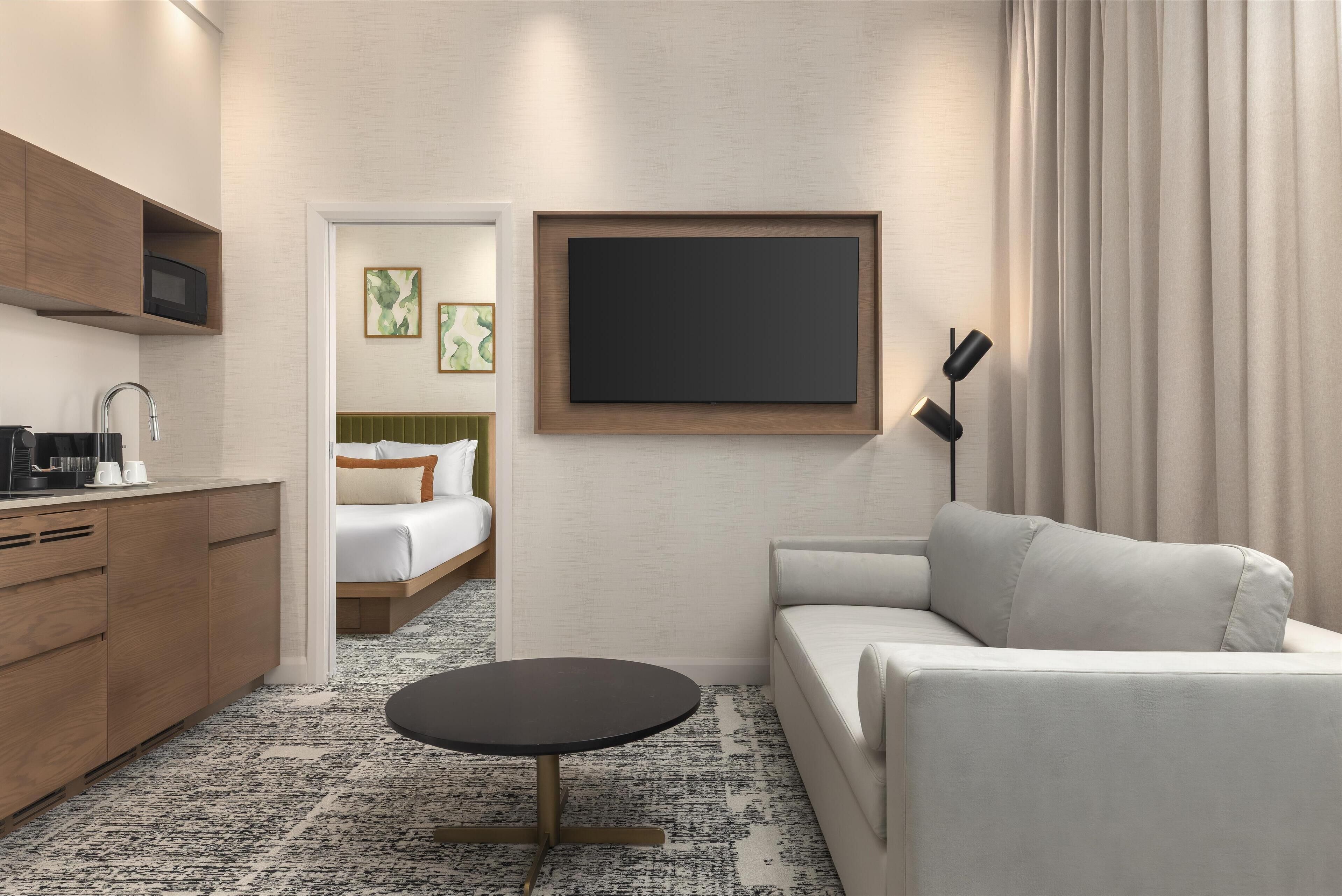 A modern hotel room with a kitchenette, a bed with a green headboard, a white sofa, a round table, and a mounted TV.