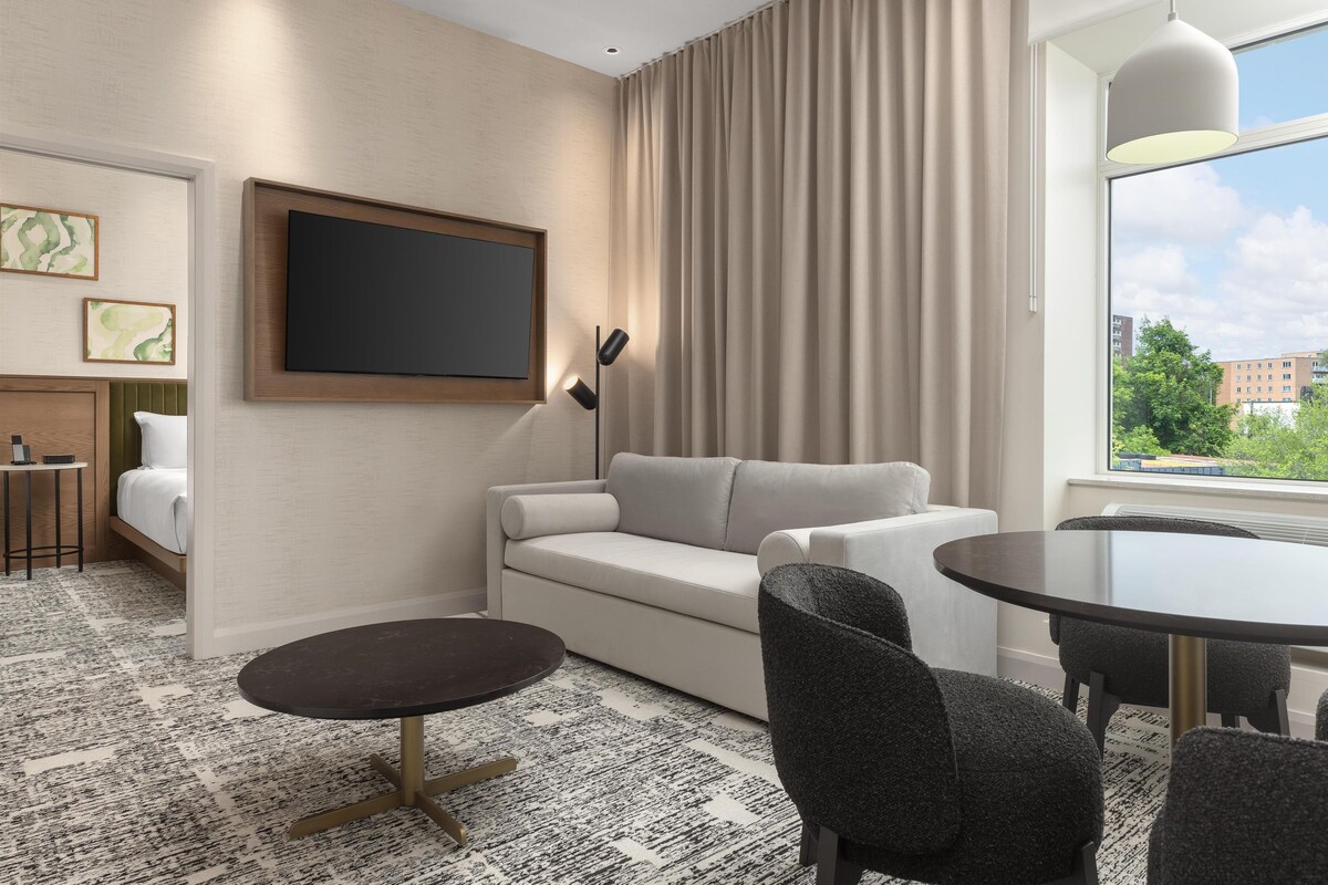 Una habitación de hotel moderna con sofá, una mesa redonda, un televisor montado en la pared y vistas de la ciudad por la ventana.