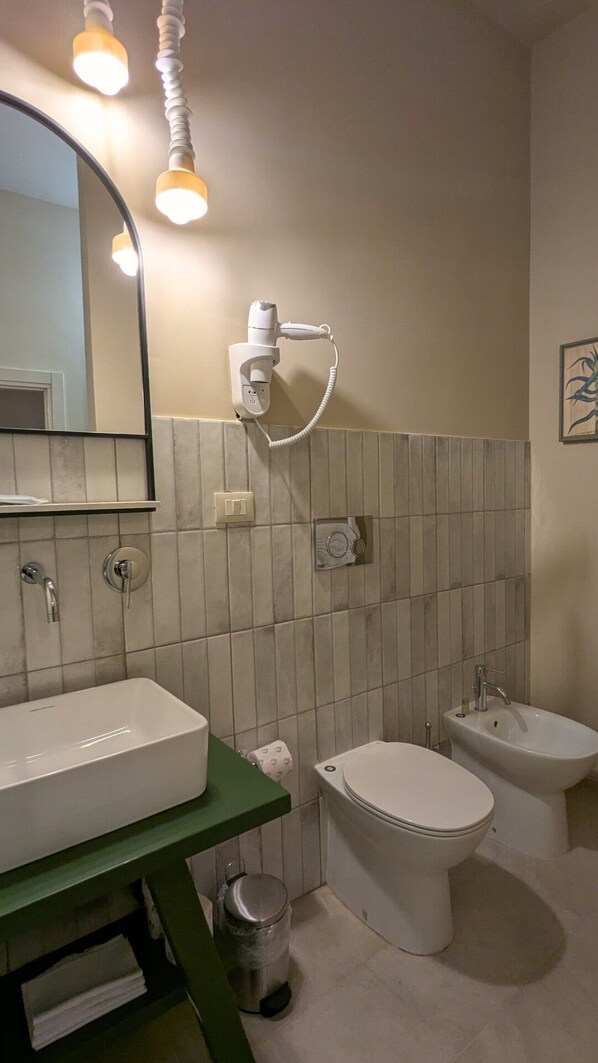 A bathroom with a white sink, a toilet, and a bidet. There is a hairdryer mounted on the wall, a mirror, and a trash can.